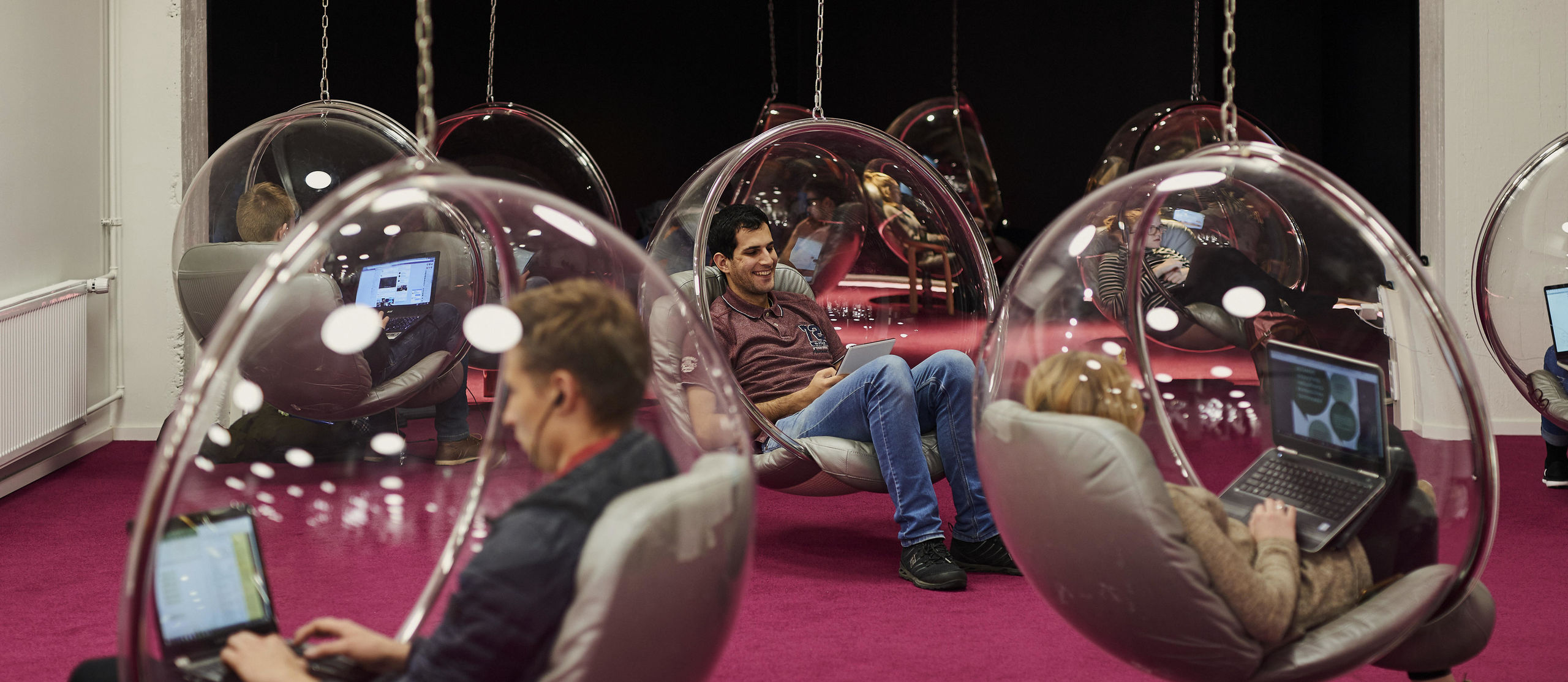 People sitting in hanging transparent chairs in a room with a pink carpet